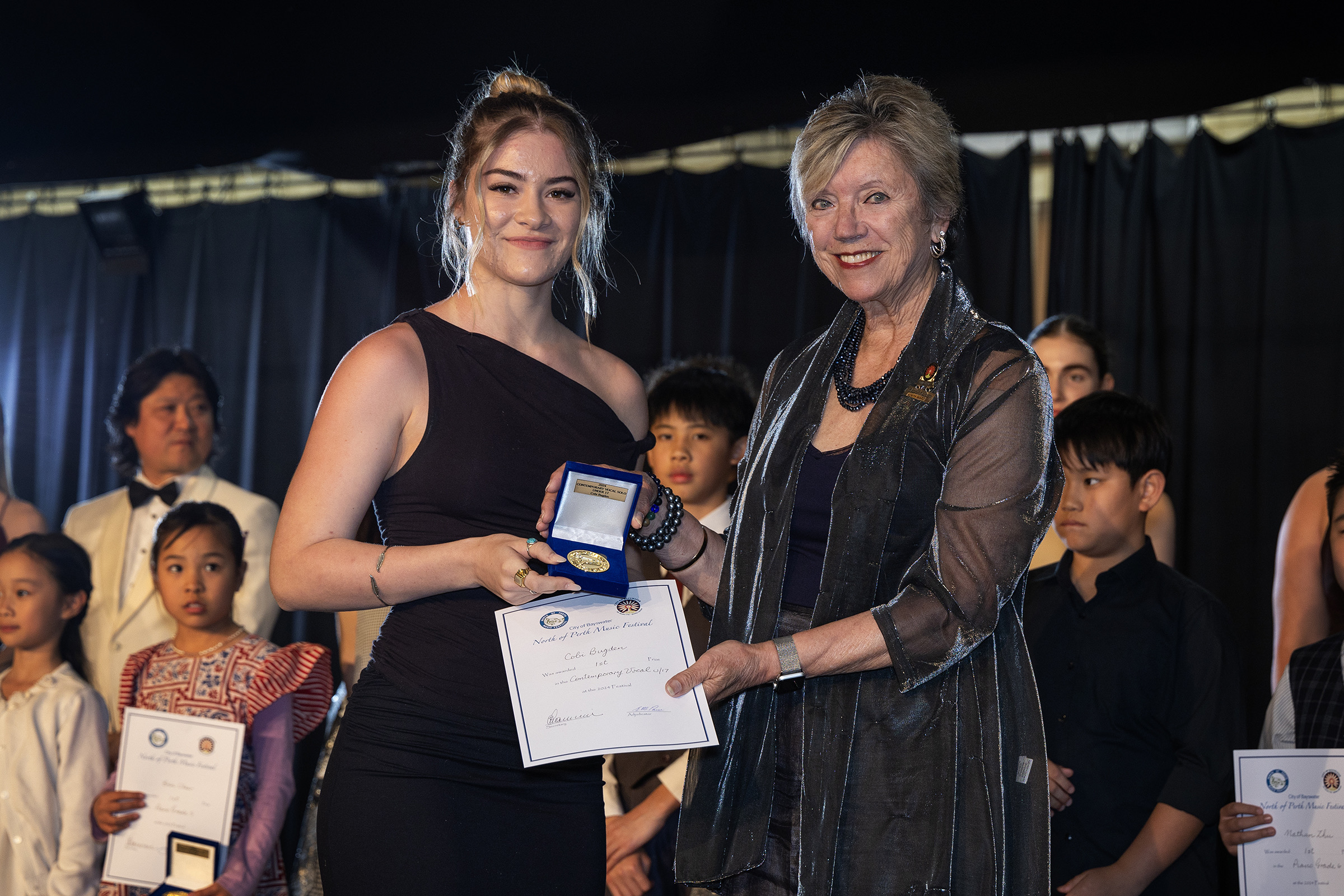 A senior winner receives her medallion and certificate from a presenter, fellow entrants looking on.