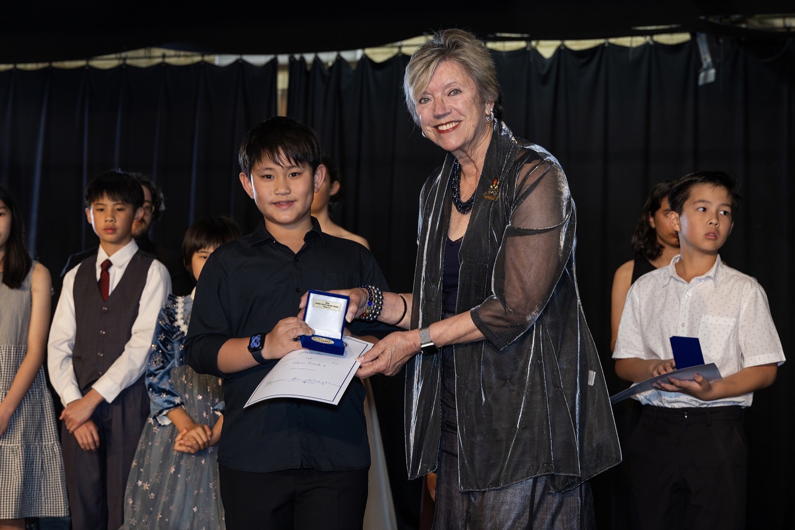 An adjudicator presents a medallion and certificate to a junior entrant, fellow entrants waiting alongside on stage.