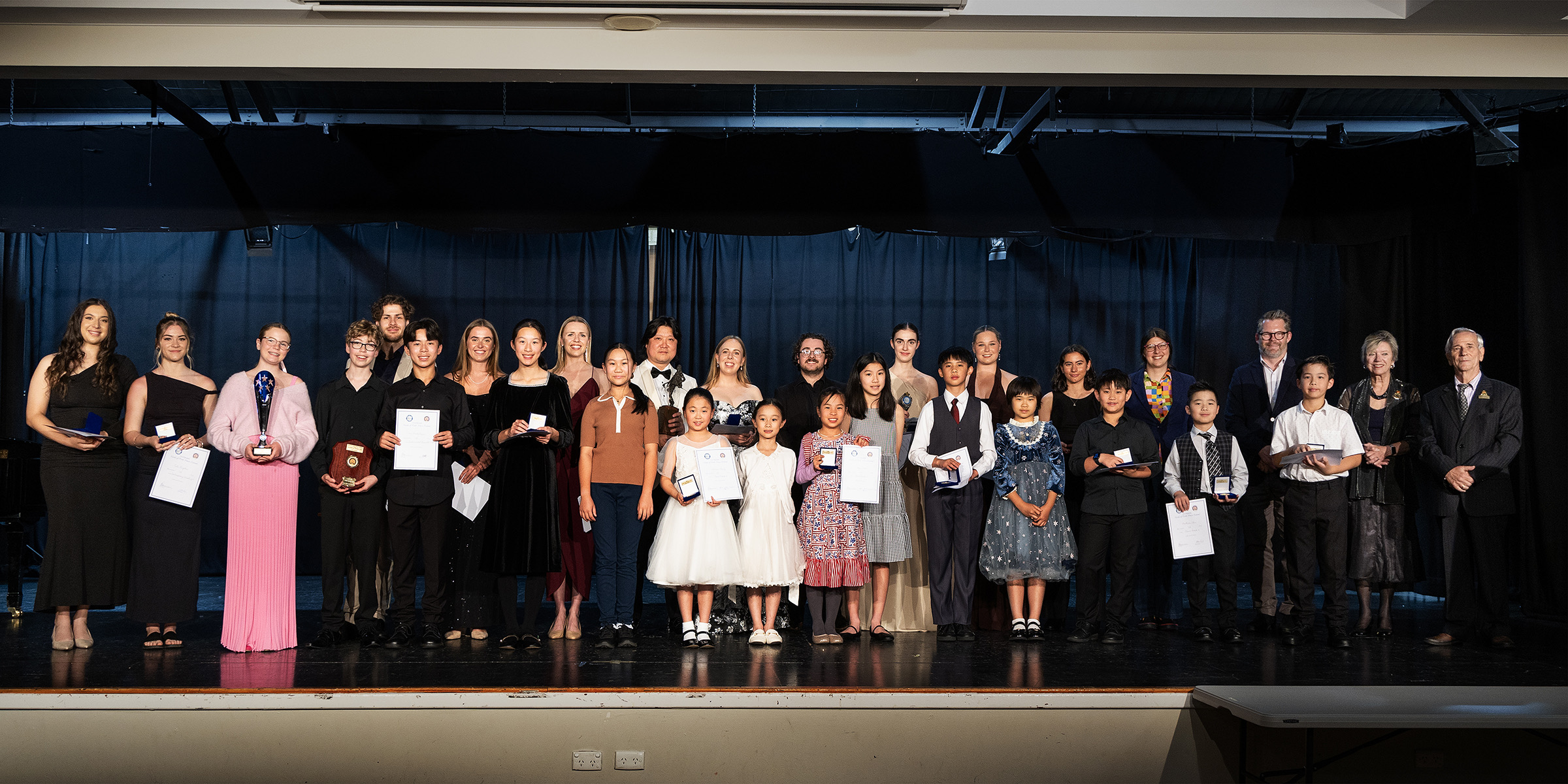 A junior winner holds a trophy, a presenter and fellow entrants beside her.