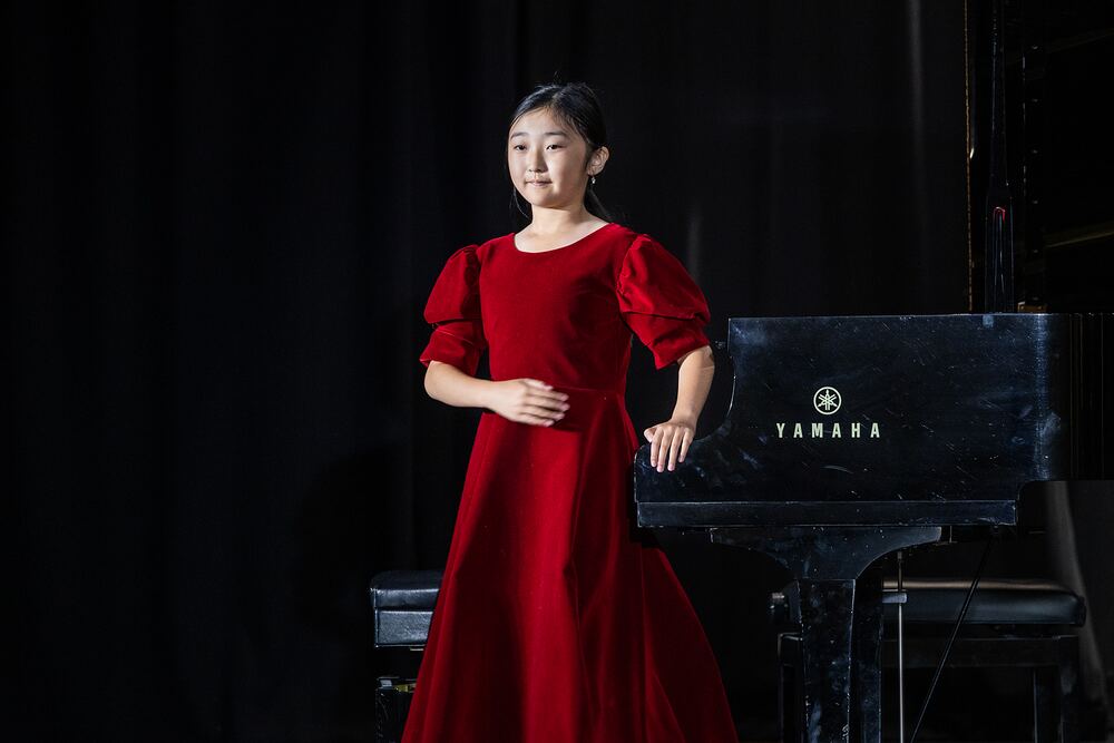 A young singer in a red velvet dress stands beside the grand piano, ready to begin.