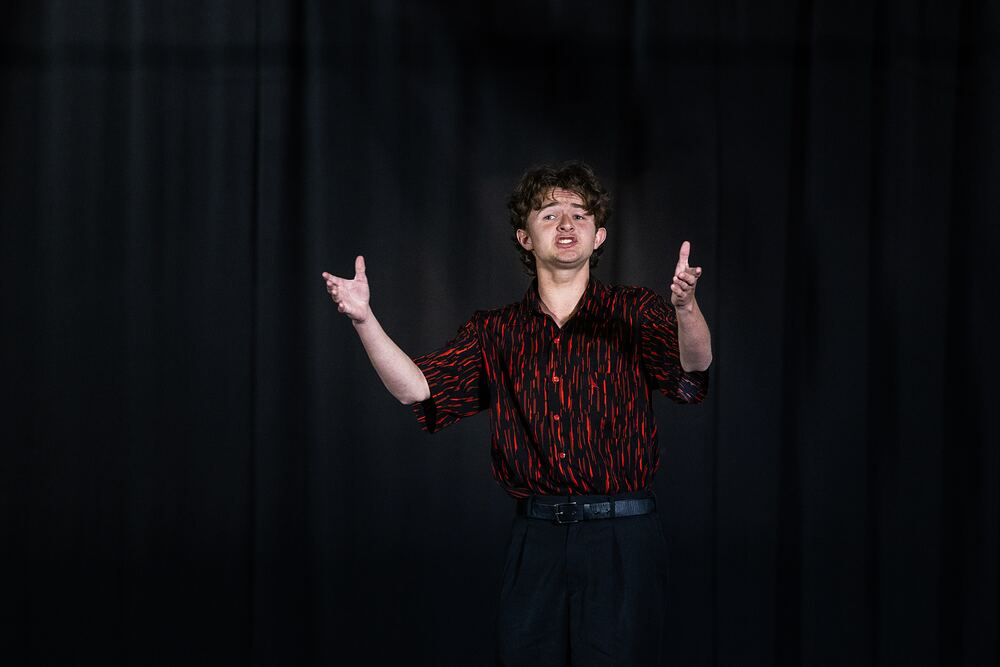 A young man in a red patterned shirt performs a speech and drama piece, arms raised.