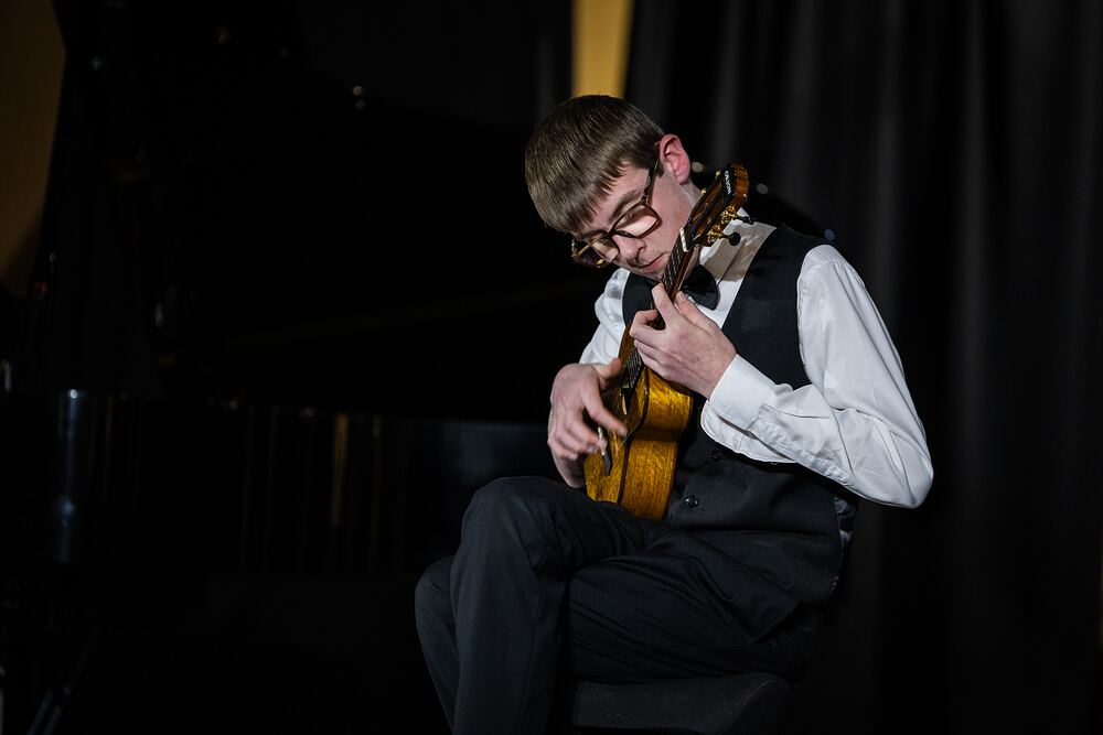 A young guitarist in a waistcoat and bow tie plays classical guitar.
