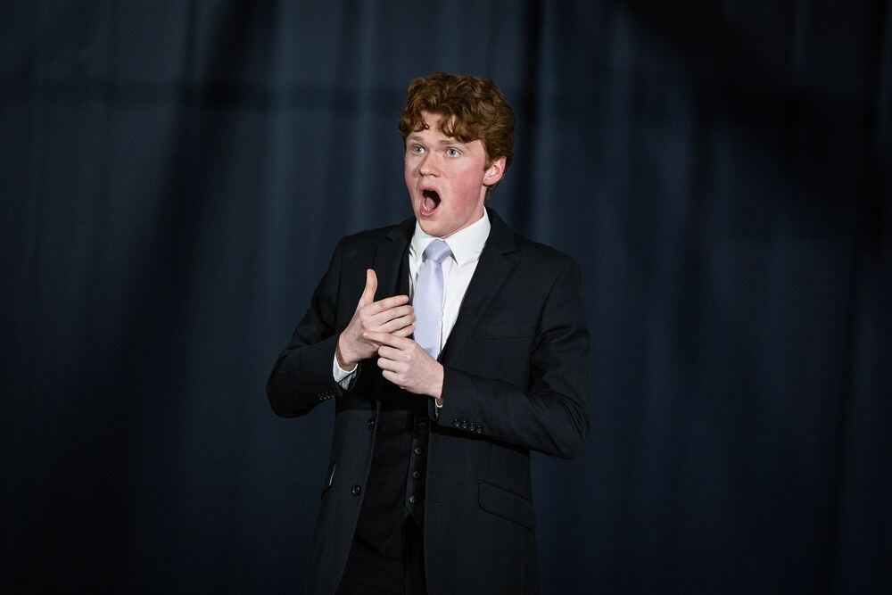 A young man in a suit performs a dramatic speech piece, hands clasped to his chest.