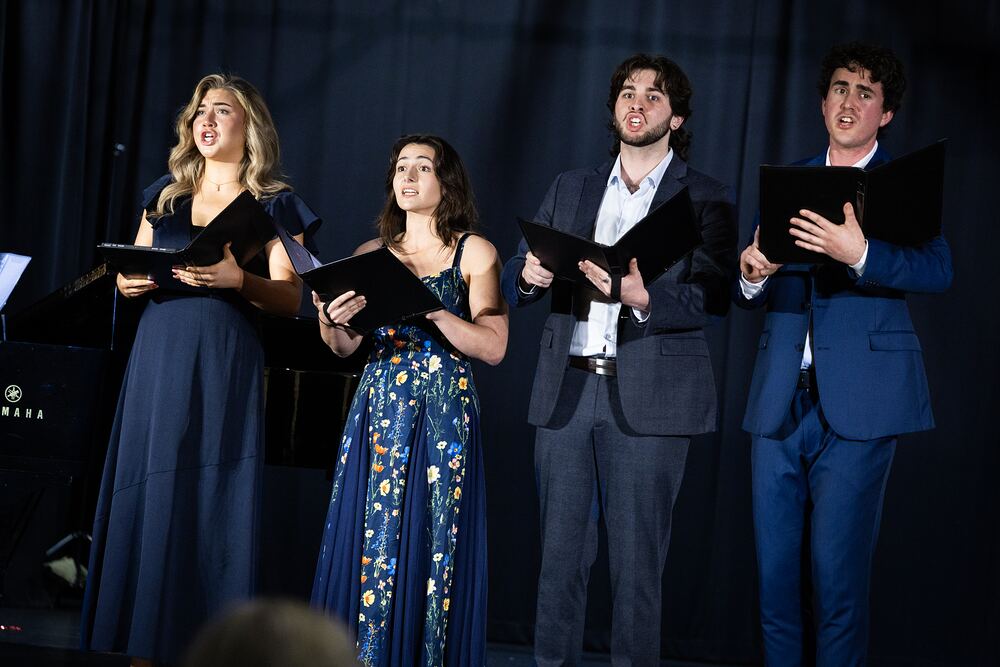 Four singers in formal wear perform together, holding black music folders.