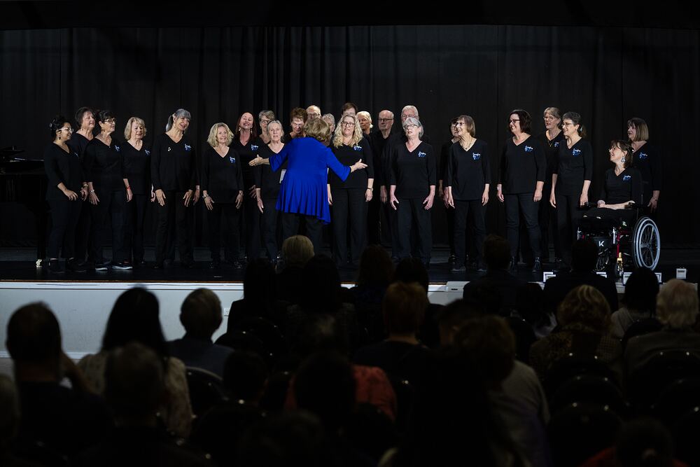 A choir dressed in black sings on stage under its conductor, the audience in the foreground.